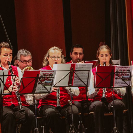 Fünf Musiker in roten Uniformen sitzen auf der Bühne und spielen Klarinette. Notenständer mit Noten sind vor ihnen. Dahinter ist ein roter Vorhang.