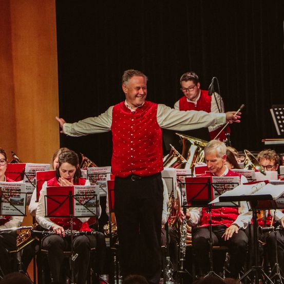 Ein Orchester tritt auf der Bühne auf, geleitet von einem Dirigenten. Die Musiker sitzen und spielen Instrumente. Der Dirigent steht mit ausgestreckten Armen. Im Hintergrund befindet sich ein dunkler Vorhang und eine Holzwand.