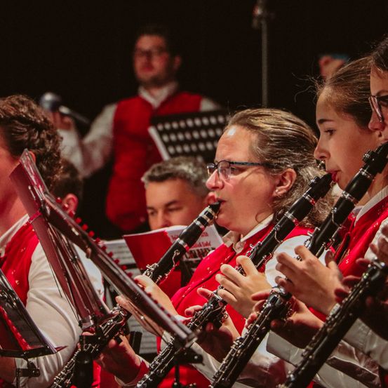 Eine Gruppe von Musikern in roten Westen spielt Klarinetten bei einer Aufführung. Ein Mann hält einen Notenständer im Hintergrund.