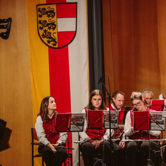 Eine Gruppe von Musikern sitzt auf Stühlen und spielt Instrumente. Sie tragen rote Westen. Hinter ihnen ist eine Fahne mit einem Wappen.