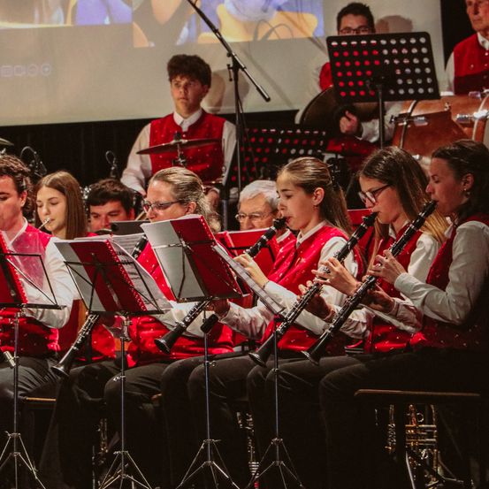 Eine Gruppe von Musikern in roten Westen spielt Instrumente, einschließlich Klarinetten, auf der Bühne. Hinter ihnen steht ein Mann mit Trommeln und Becken. Eine Leinwand projiziert ein Bild im Hintergrund.