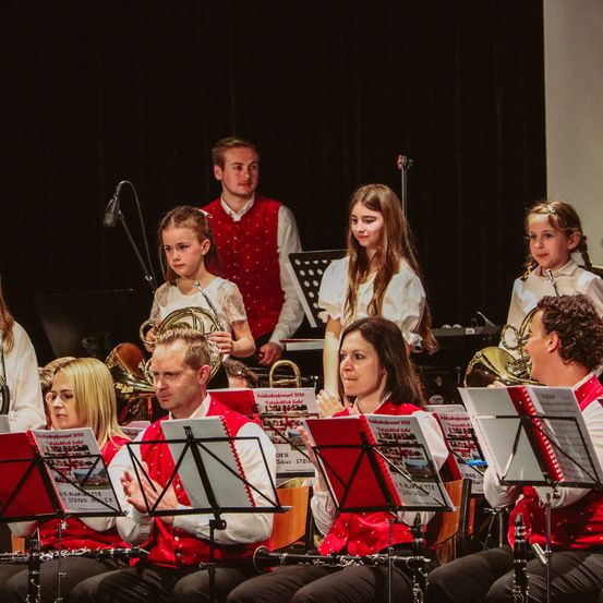 Eine Gruppe von Musikern tritt auf der Bühne auf. Einige stehen und andere sitzen mit Instrumenten vor ihnen. Hinter ihnen ist ein schwarzer Vorhang.