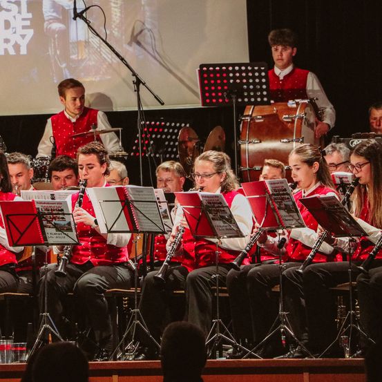 Eine Schulband spielt auf der Bühne, mit einer Projektionsleinwand im Hintergrund. Mehrere Musiker spielen Instrumente, einschließlich Trommeln und Klarinetten.