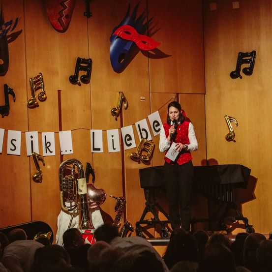 Eine Frau steht mit einem Mikrofon auf der Bühne vor einer Kulisse aus Musiknoten und Masken. Hinter ihr stehen Musikinstrumente und eine Menge schaut zu.