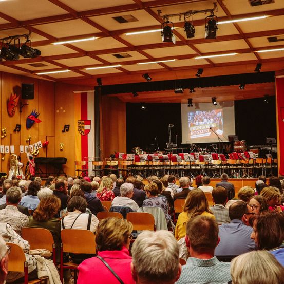 Ein Saal voller Menschen, die ein Konzert sehen. Die Bühne hat Musikinstrumente und eine große Leinwand mit Text. Musiknoten und Flaggen sind an der Wand montiert.