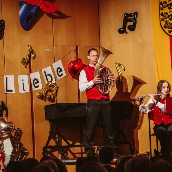 Ein Mann und eine Frau spielen auf der Bühne mit Musikinstrumenten vor einem Publikum. Der Mann spielt Tuba, die Frau Flöte. Hinter ihnen ist eine Wand, die mit Noten und einem Banner verziert ist, das 'Liebe' besagt.