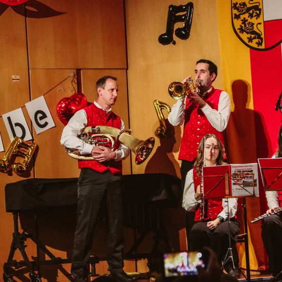 Eine Gruppe von Musikern tritt auf einer Bühne mit roten und gelben Kulissen auf. Ein Mann spielt Trompete, ein anderer Tuba und eine Frau Flöte.