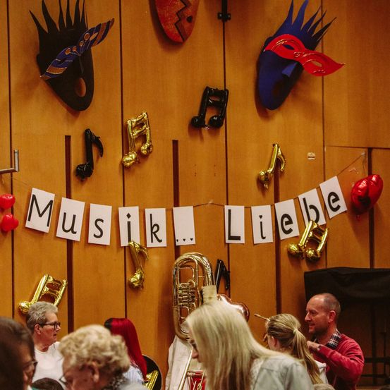 Ein Raum mit Musikinstrumenten und einem Banner mit der Aufschrift 'Musik...Liebe'. Menschen stehen und beobachten. Musiknoten und Masken sind an der Wand angebracht.