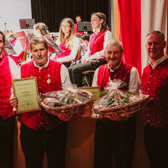 Vier Männer in roten Westen stehen stolz und halten Blumenkörbe. Ein Mann in der Mitte hält eine Auszeichnung. Hinter ihnen spielt eine Gruppe Musik mit Instrumenten.