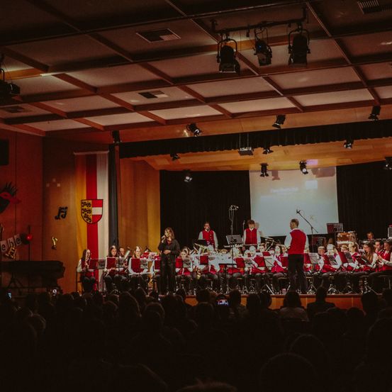 Ein großer Innenraum mit Musikern, die auf der Bühne vor einem großen Publikum auftreten. Sie haben verschiedene Instrumente, darunter Trompeten, Posaunen und Trommeln.