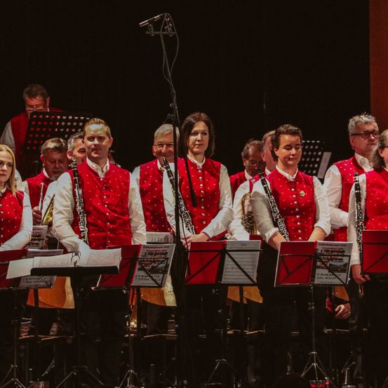 Eine Gruppe von Musikern in roten Westen steht vor Notenständern und hält Musikinstrumente wie Klarinetten und Trompeten. Sie scheinen sich auf eine Aufführung auf der Bühne vorzubereiten.