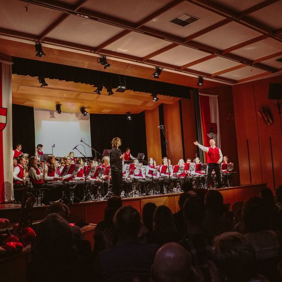 Eine Band spielt auf der Bühne mit Fahnen im Hintergrund, unter Scheinwerfern. Zuschauer sitzen im Vordergrund.