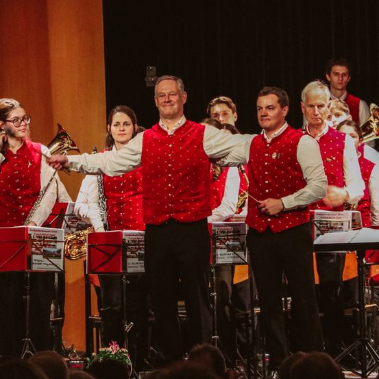 Ein Orchester spielt auf der Bühne, angeführt von einem Dirigenten. Die Orchestermitglieder tragen rote Westen mit weißen Punkten. Sie spielen verschiedene Musikinstrumente wie Trompeten und Posaunen. Vor ihnen stehen Notenständer mit Musikblättern.