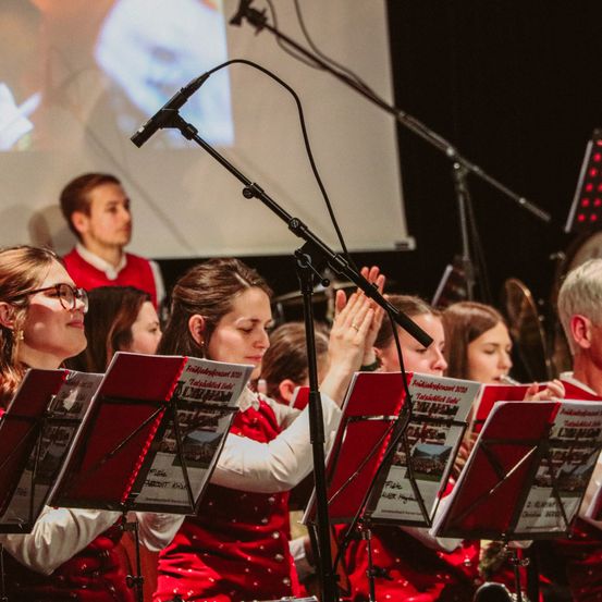 Eine Band in roten Uniformen tritt auf der Bühne auf, mit einem Mikrofon im Vordergrund. Ein Projektionsbildschirm ist hinter ihnen zu sehen.