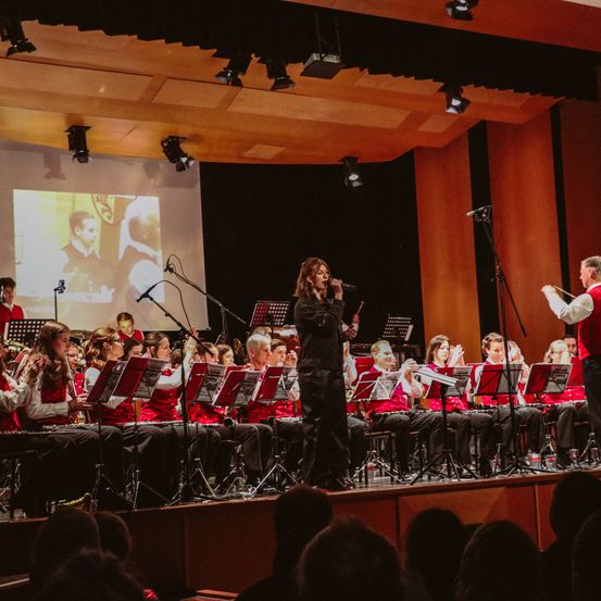 Eine Frau singt auf der Bühne, während eine große Band hinter ihr spielt. Das Publikum sitzt vor ihnen. Eine Projektionsleinwand zeigt einen Mann im Hintergrund.