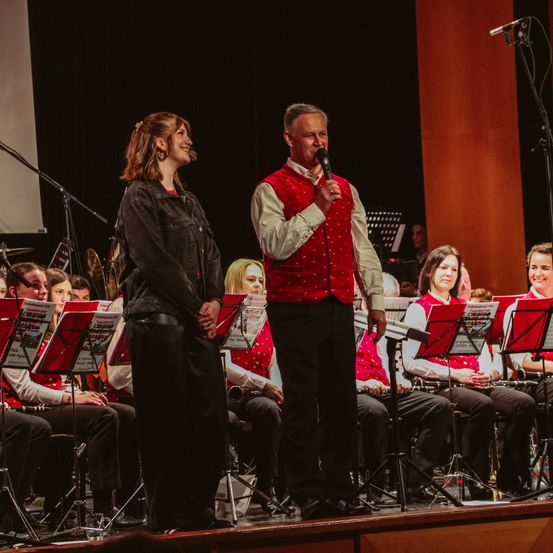 Ein Mann und eine Frau stehen auf der Bühne mit Musikinstrumenten im Hintergrund. Der Mann hält ein Mikrofon und scheint zu singen. Das Publikum sitzt und hört aufmerksam zu.