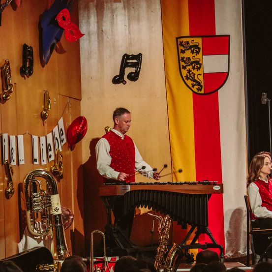 Ein Mann spielt ein Xylophon auf der Bühne, mit einer Flagge und Noten im Hintergrund. Eine Frau sitzt vor ihm in einem Stuhl. Verschiedene Instrumente sind an der Wand ausgestellt.