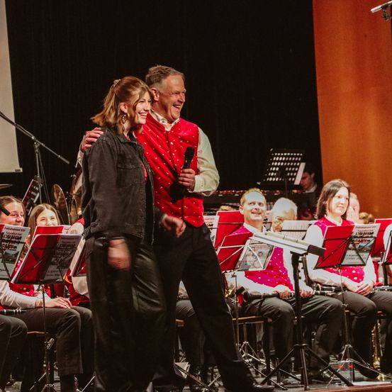 Eine Gruppe von Musikern auf der Bühne mit einer Frau und einem Mann, die lächelnd vor ihnen stehen. Der Mann hält ein Mikrofon und scheint zu sprechen.