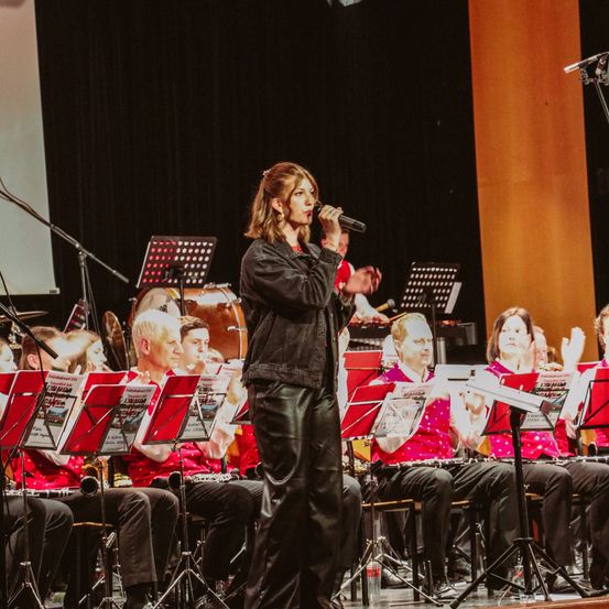 Eine Sängerin steht auf der Bühne mit einer Band und hält ein Mikrofon. Hinter ihr spielen Musiker verschiedene Instrumente, während das Publikum jubelt. Die Bühne ist mit Notenständern und Instrumenten ausgestattet.