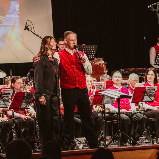Ein Mann und eine Frau stehen auf der Bühne vor einer Band. Der Mann hält ein Mikrofon, während die Frau lächelt. Hinter ihnen sitzt die Band mit Musikinstrumenten und Notenblättern.
