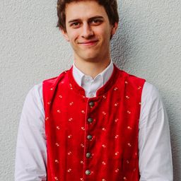 Ein lächelnder junger Mann posiert vor einer Wand in einem roten Westchen mit Blumenmuster und einem weißen Hemd.