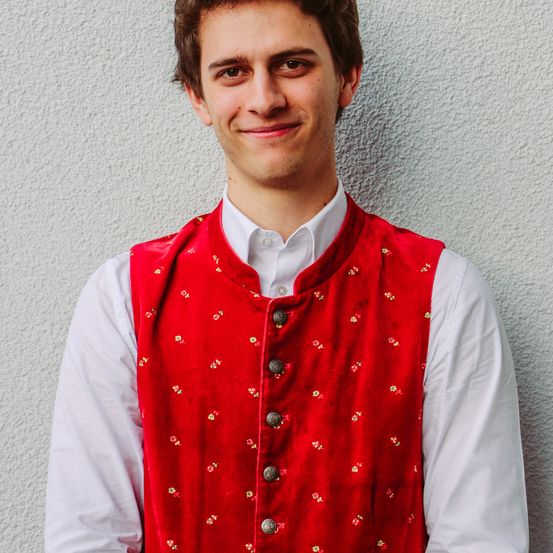 Ein lächelnder junger Mann posiert vor einer Wand in einem roten Westchen mit Blumenmuster und einem weißen Hemd.