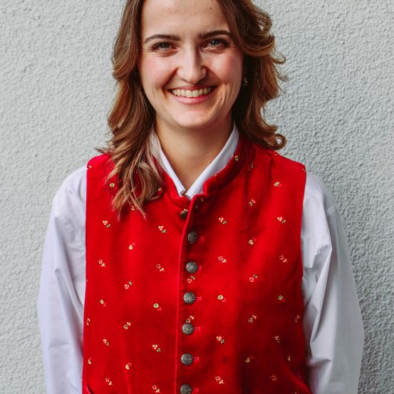 Eine lächelnde Frau mit lockigen Haaren trägt eine rote Weste mit Blumenmuster und ein weißes Hemd. Sie steht vor einer weißen Wand.