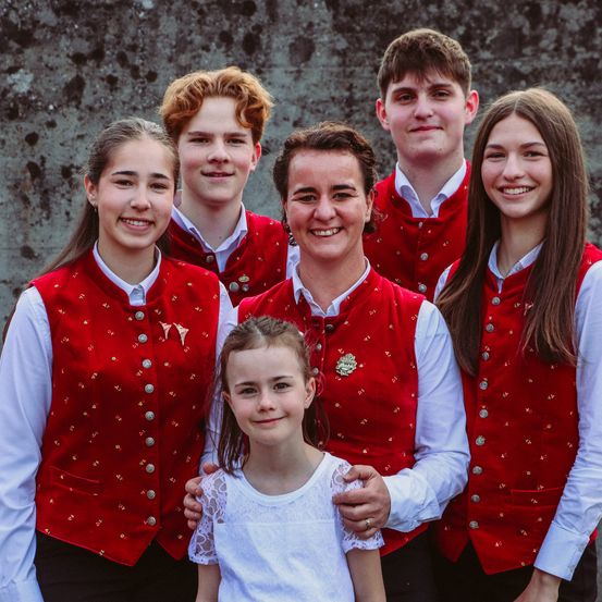 Eine Familie von sechs Personen, alle in übereinstimmenden roten Westen gekleidet, posiert für ein Foto vor einem Steinmauerhintergrund. Ein junges Mädchen in einem weißen Kleid steht vor einer Frau und einem Jungen.