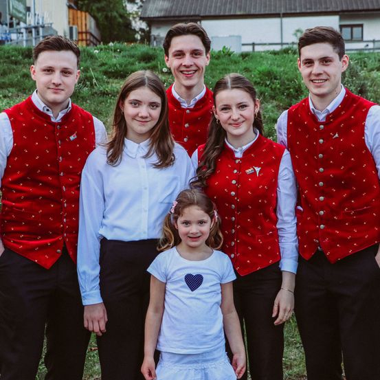 Eine Gruppe von Menschen, darunter vier junge Männer und drei Mädchen, stehen auf einem Feld, lächeln und posieren für ein Foto. Sie tragen alle rote Westen und schwarze Hosen.