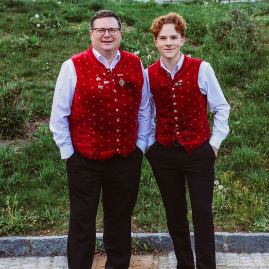 Zwei Männer stehen nebeneinander in roten Westen und schwarzen Hosen auf einem Bürgersteig. Sie lächeln und posieren für ein Foto vor einem grünen Hintergrund.