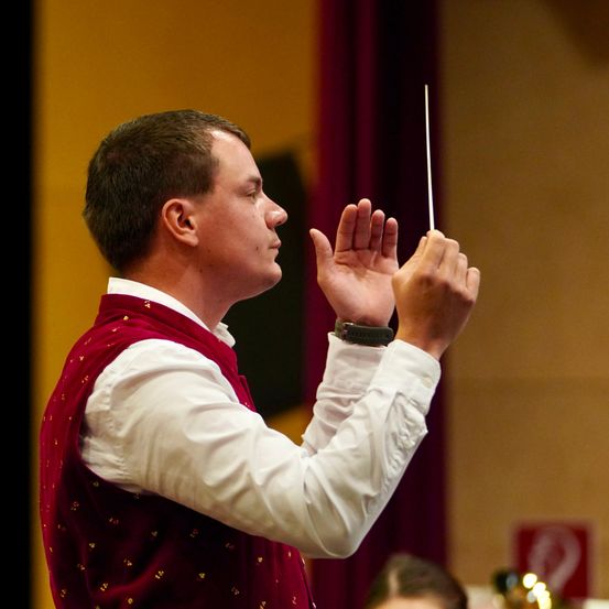 Ein Mann in einem roten Weste mit goldenen Mustern dirigiert ein Orchester mit einem weißen Dirigentenstab in einem Konzertsaal.