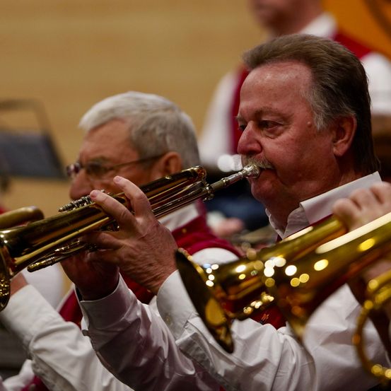 Zwei Männer spielen Blechblasinstrumente in einem Konzertsaal. Der Mann im Vordergrund trägt eine rote Weste und ein weißes Hemd, während der Hintergrund einen anderen Musiker zeigt.