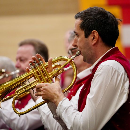 Ein Trompeter in einem roten Vest konzentriert sich auf seine Darbietung, umgeben von anderen Musikern. Der Schauplatz ist ein Konzert.