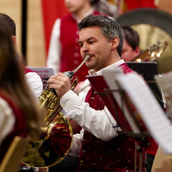 Ein Mann in einer roten Weste spielt ein Blechinstrument in einem Musikensemble. Der Hintergrund deutet auf eine formelle Veranstaltung mit anderen Musikern hin.
