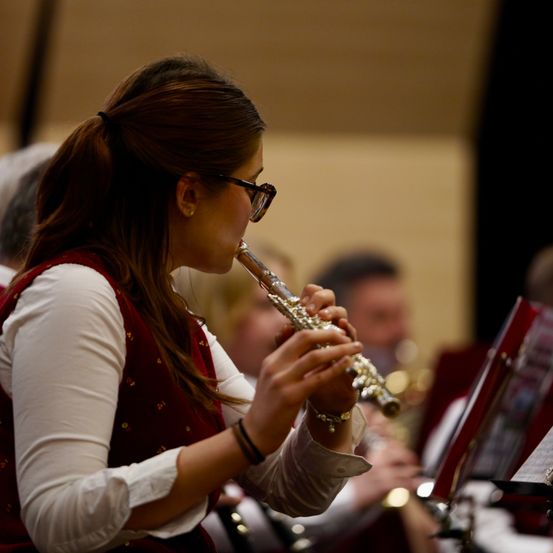 Eine Frau mit Brille und einem roten Westchen spielt Flöte in einer Band. Andere Bandmitglieder spielen im Hintergrund ihre Instrumente.