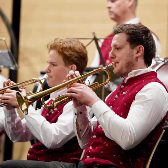 Zwei junge Männer spielen Trompeten bei einer musikalischen Aufführung. Sie tragen rote Westen mit goldenen Akzenten und sitzen auf Stühlen. Ein weiterer Musiker steht hinter ihnen und schaut sie an.