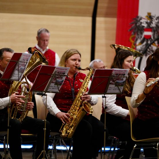 Ein Blasorchester spielt, wobei eine Frau die Posaune und andere die Trompete spielen. Sie sitzen auf Stühlen und sind dem Publikum zugewandt, mit Notenständern vor ihnen.