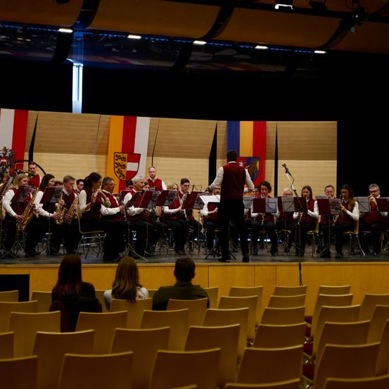 Ein Orchester spielt auf der Bühne mit Instrumenten. Die Bühne ist mit Flaggen geschmückt. Das Publikum sitzt auf Stühlen und beobachtet die Aufführung.