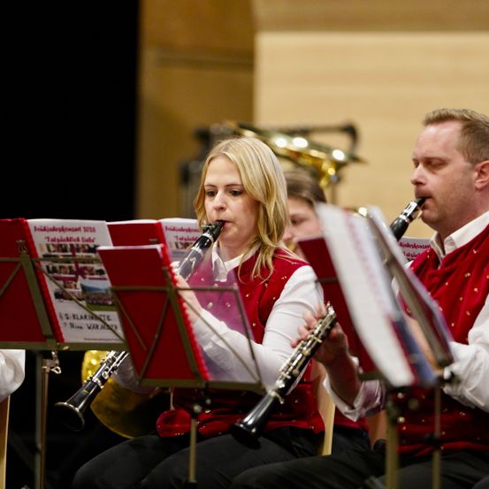 Zwei Musiker, ein Mann und eine Frau, sitzen auf der Bühne und spielen Klarinetten. Sie sind in roten Westen und weißen Hemden gekleidet. Die Frau hat blondes Haar. Hinter ihnen befinden sich rote Notenständer.