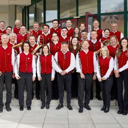A group of musicians in red vests pose for a photo outside a building with large windows.