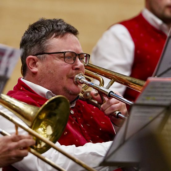 Ein Mann in einer roten Weste und mit einer Brille spielt während eines Konzerts eine Posaune, mit einem anderen Musiker im Hintergrund.