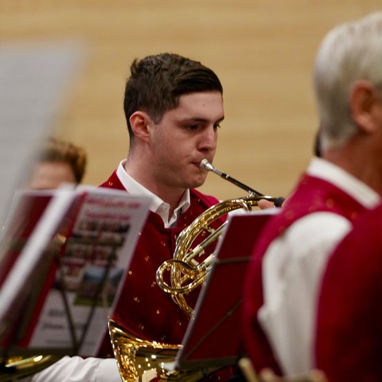 Ein junger Mann in einem roten Kostüm spielt während einer Bandaufführung eine französische Horn. Er ist konzentriert, hält das Instrument mit beiden Händen, während ein anderer Bandmitglied hinter ihm steht.