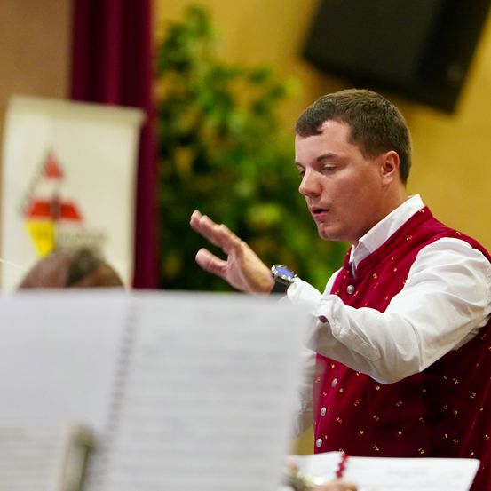 Ein Mann in einem roten Weste dirigiert ein Musikensemble, seine Hände bewegt, konzentriert auf die Noten vor ihm.