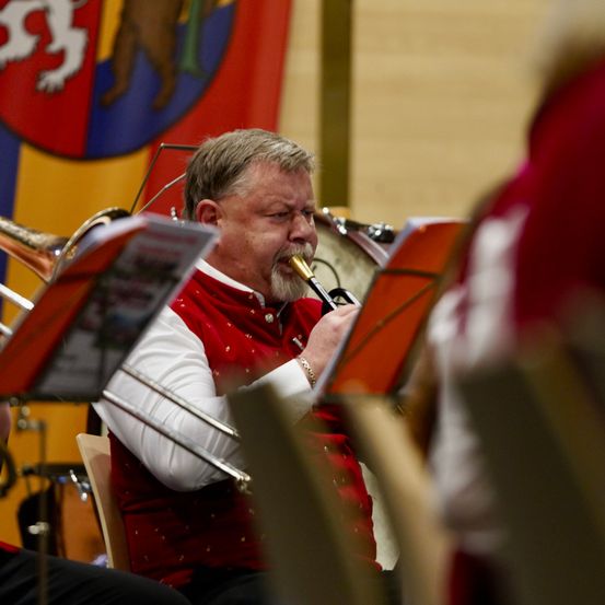 Ein Mann in einer roten Weste spielt bei einem Konzert ein Blechblasinstrument. Hinter ihm ist eine Fahne.