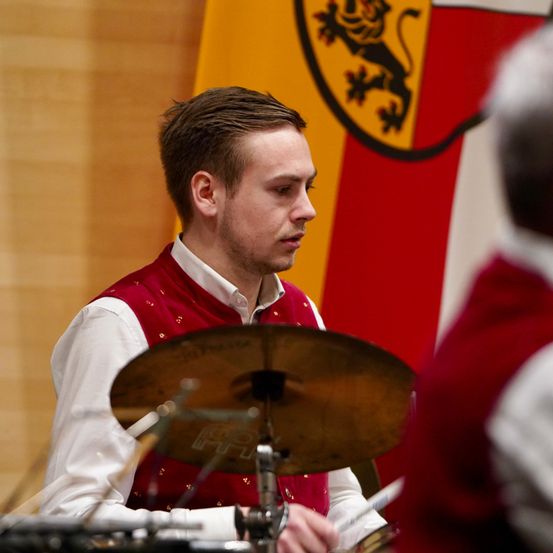 Ein Mann in einer roten Weste und weißem Hemd spielt Schlagzeug mit einer Becken vor einer Fahne mit Löwenwappen.