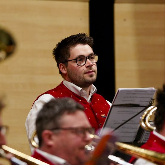 Ein Mann in einer roten Weste und mit Brille liest Musik und hält ein Posaune. Andere Musiker sind hinter ihm.