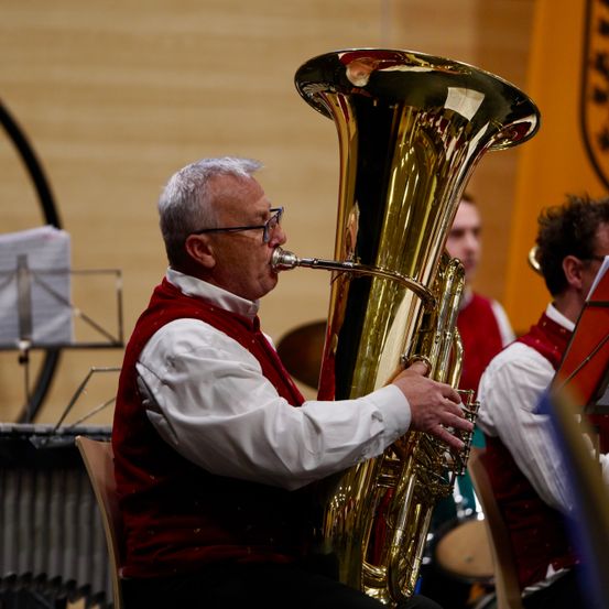 Ein älterer Mann spielt eine Tuba in einem musikalischen Ensemble. Er trägt eine rote Weste und Brille und konzentriert sich auf sein Instrument. Hinter ihm spielt ein weiterer Musiker und erzeugt einen harmonischen Klang.