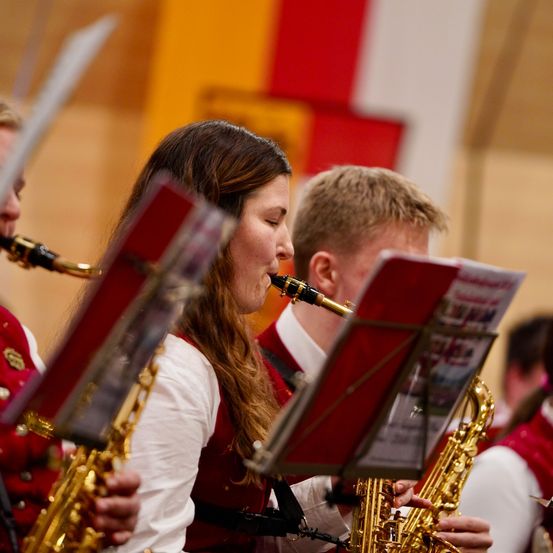 Eine Saxophonband spielt, wobei eine Frau vorne ein Saxophon spielt und ein Mann daneben ebenfalls ein Saxophon spielt. Beide tragen rote Westen.