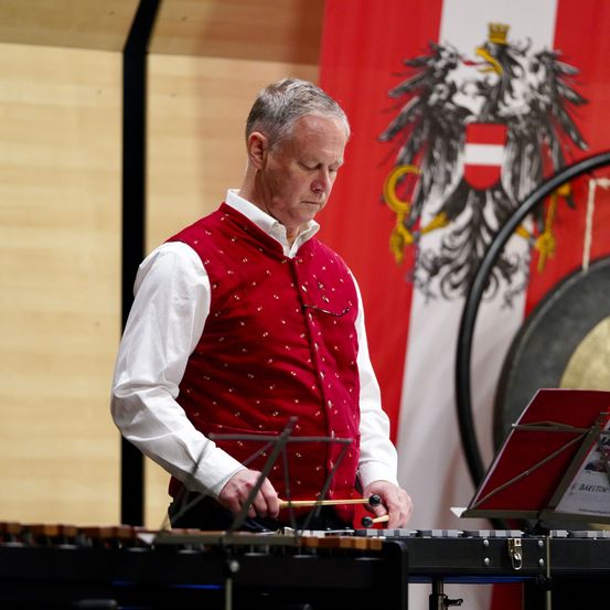 Ein älterer Mann spielt ein Xylophon auf der Bühne. Er trägt eine rote Weste und ein weißes Hemd. Hinter ihm befindet sich eine Flagge mit einem großen Adler und einem roten Schild.