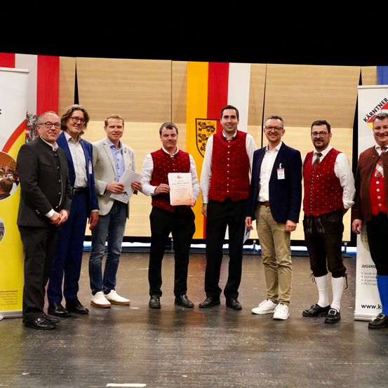 Eine Gruppe von Männern in traditioneller und formaler Kleidung steht auf der Bühne und hält ein Zertifikat in der Hand. Sie lächeln für die Kamera.